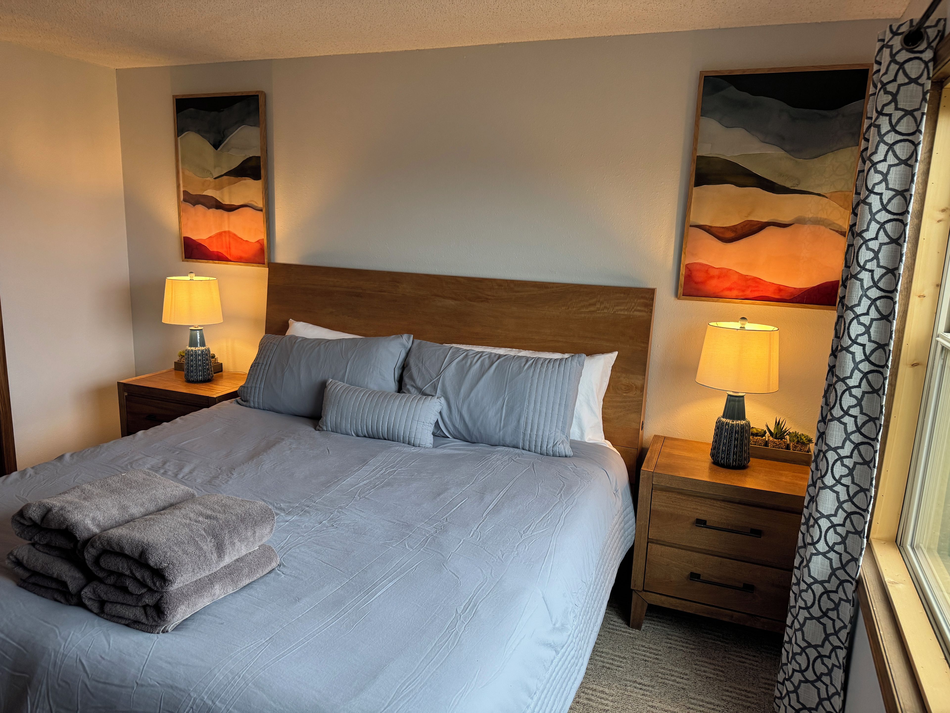 A neatly made bed with pillows and a folded blanket, two bedside tables with lamps, and two wall-mounted paintings.