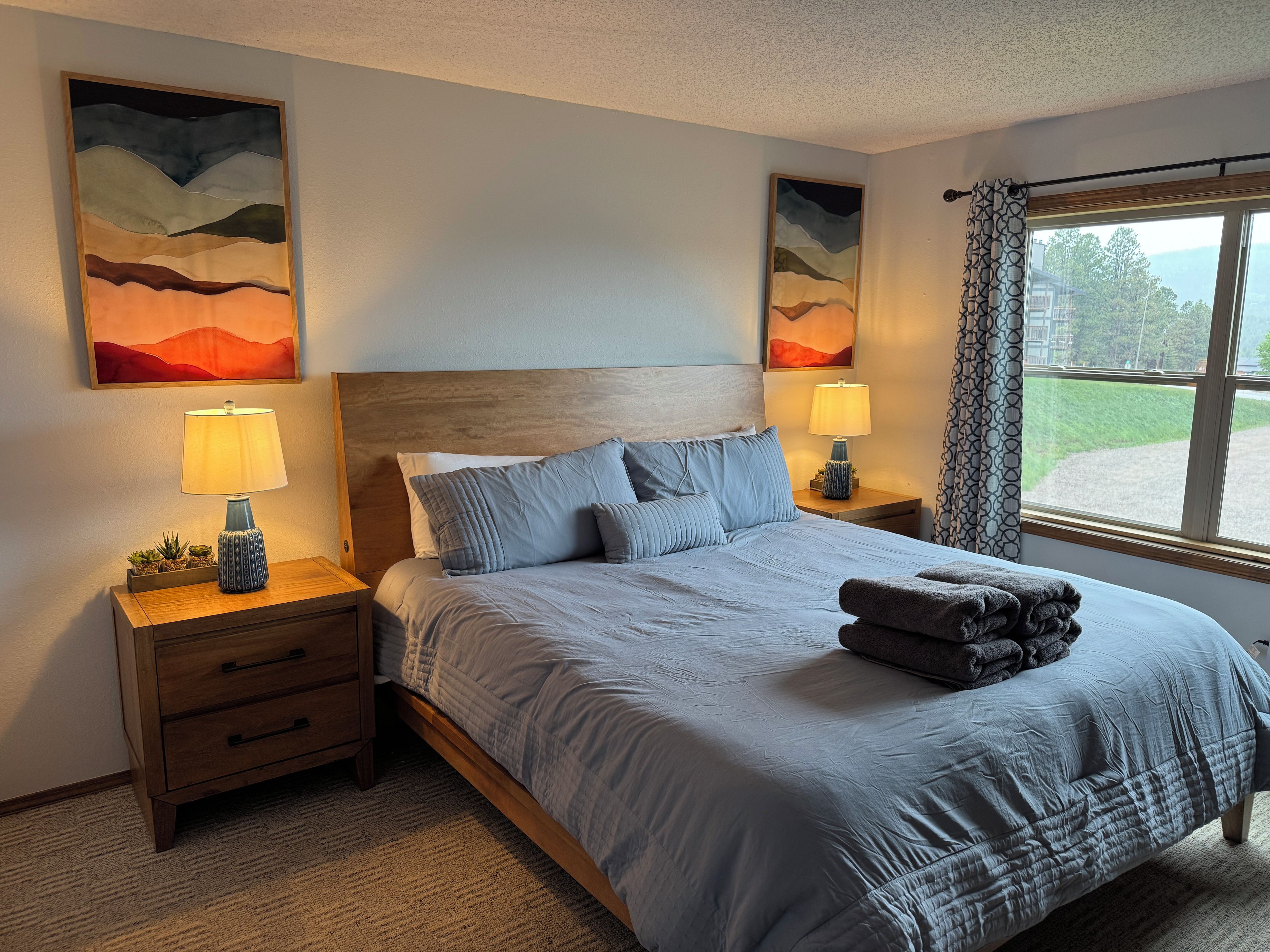 A bedroom with a bed, two bedside tables with lamps, a wooden headboard, and artwork on the walls.