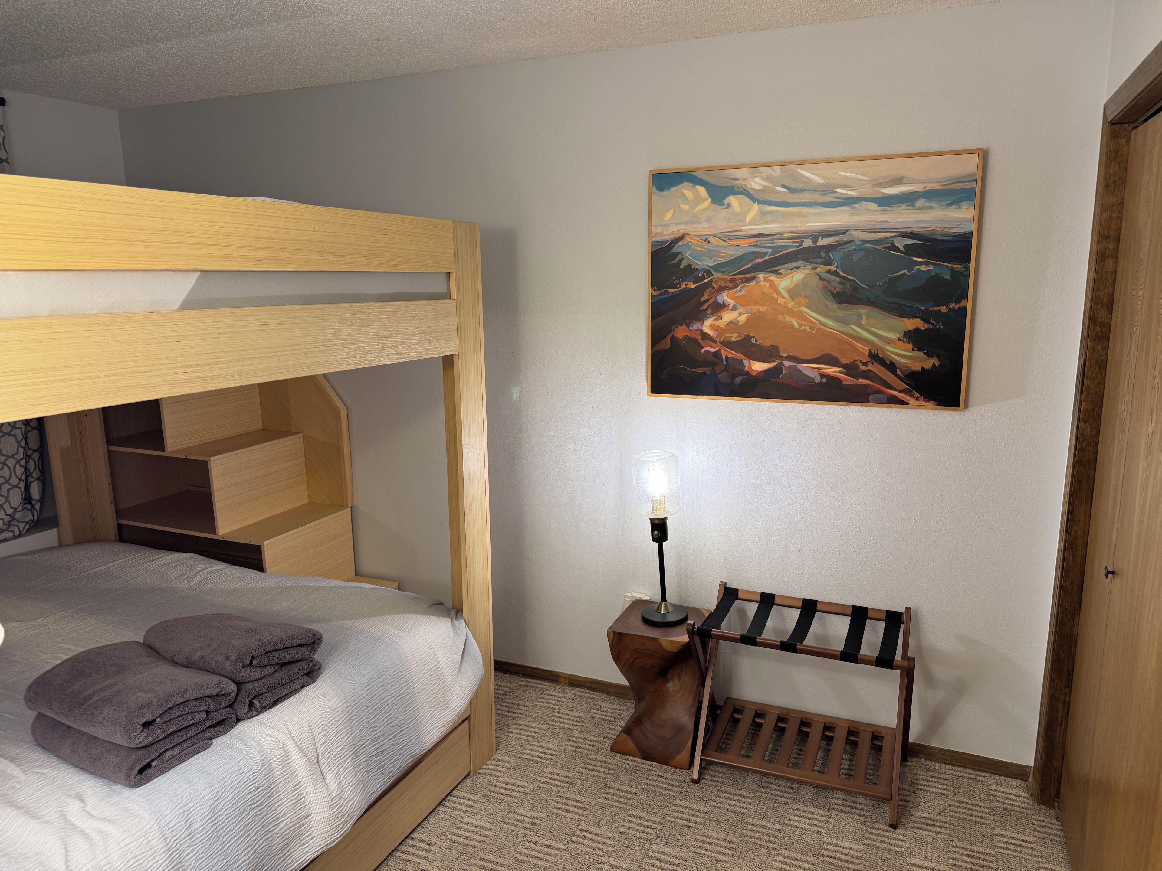 A bunk bed with a headboard, a bedside table with a lamp, a bench, and a painting on the wall.
