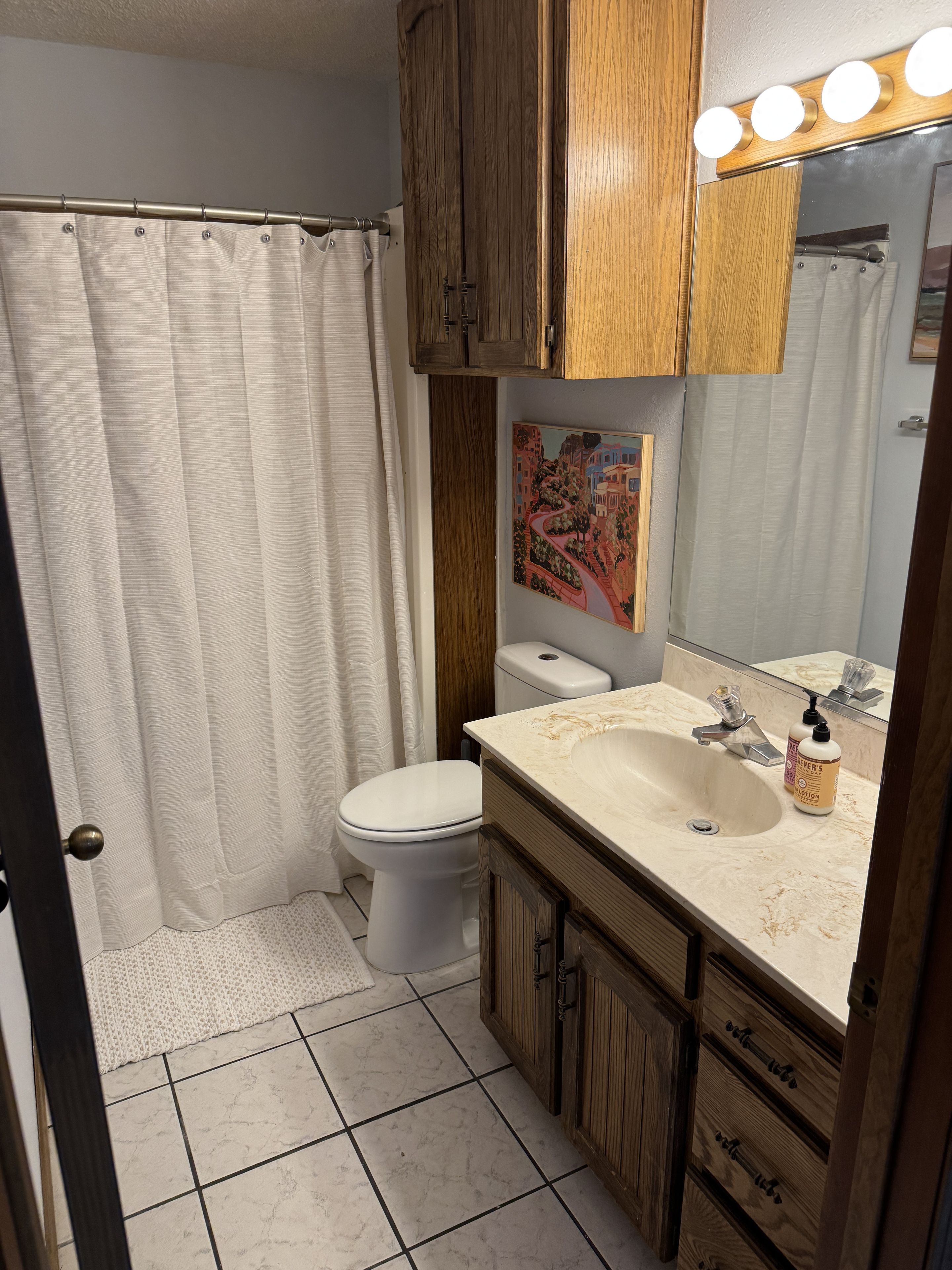 A bathroom with a toilet, sink, wooden cabinets, a mirror with lights, and a shower curtain.