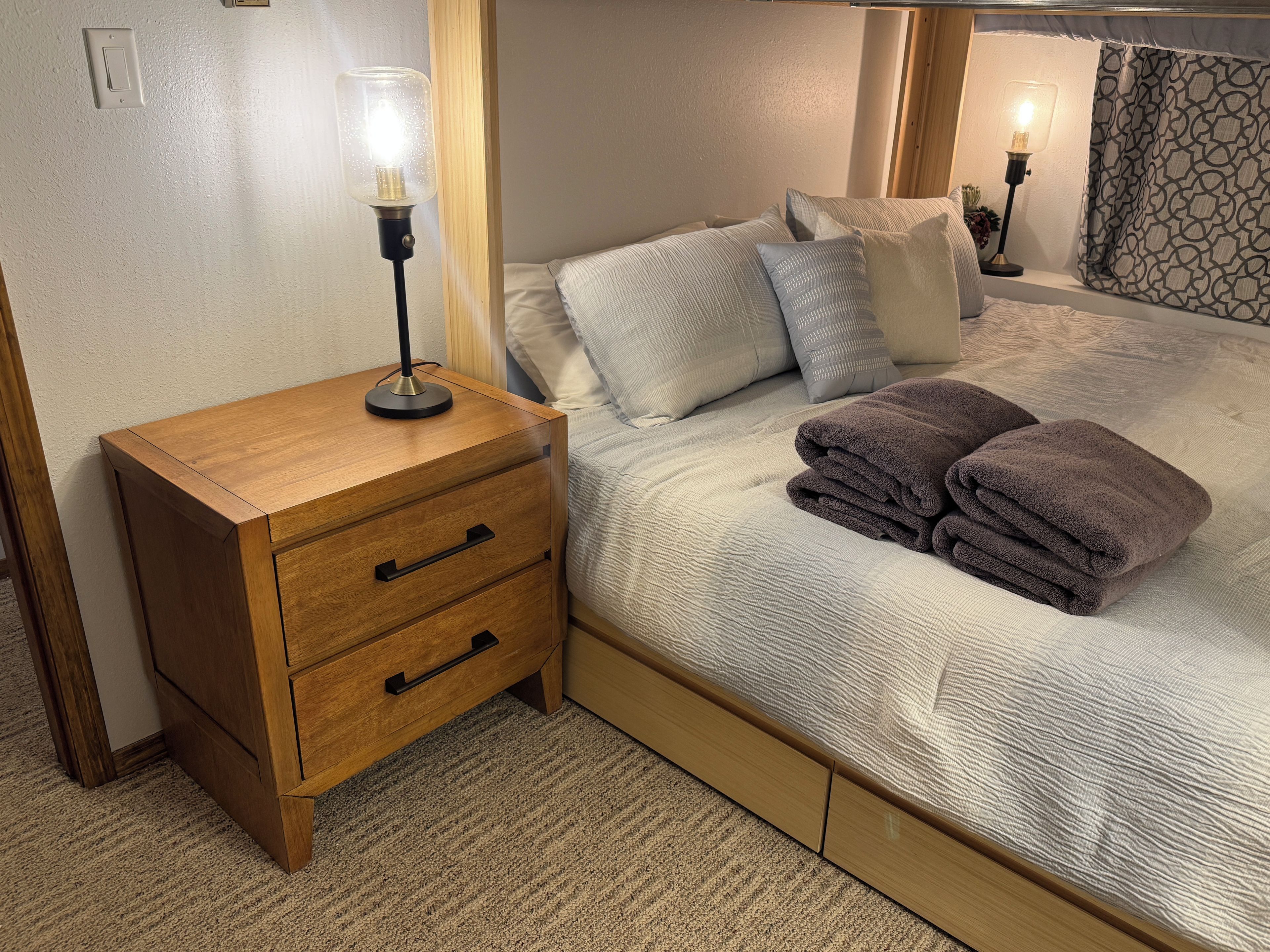 A neatly made bed with pillows and a folded towel, a wooden bedside table with a lamp, and a carpeted floor.