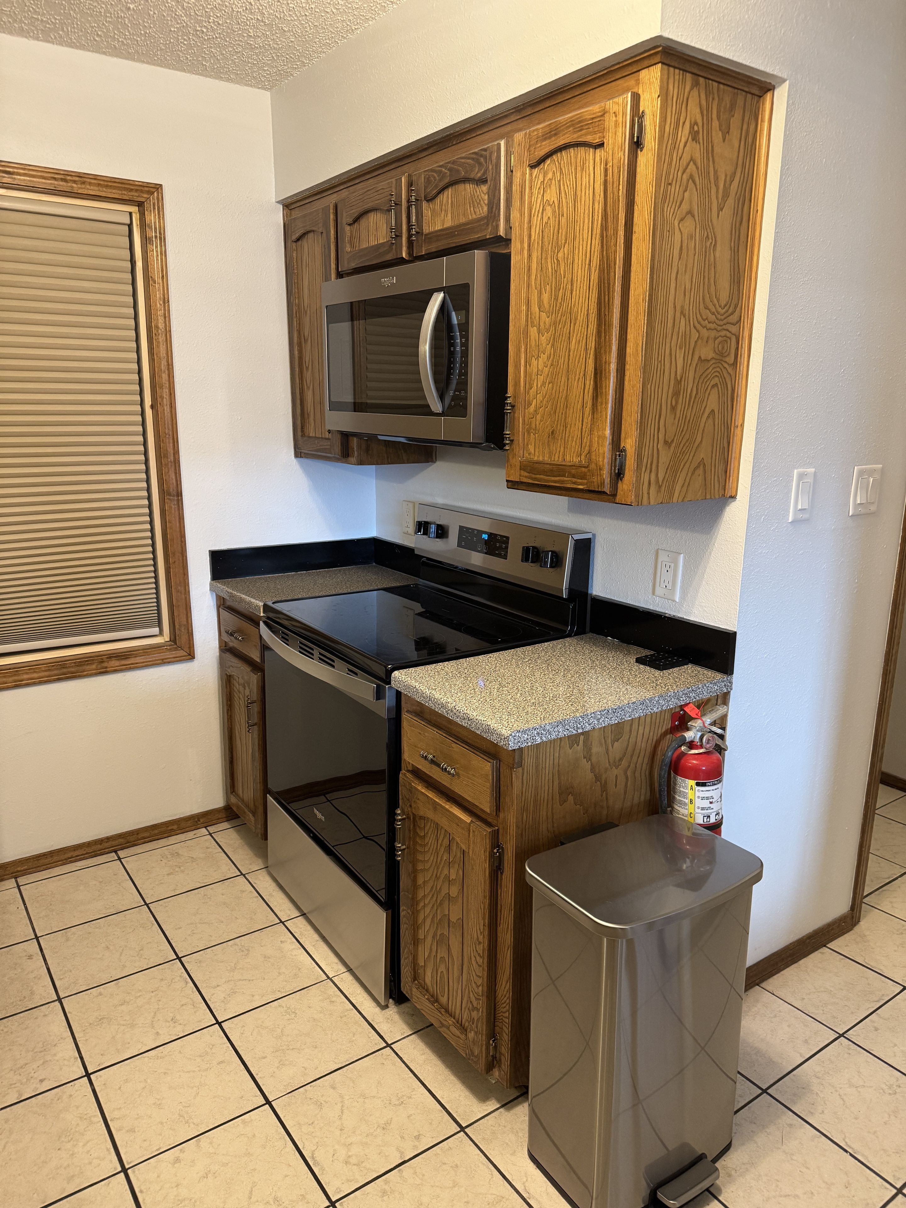 A kitchen with wooden cabinets, a stainless steel microwave, a stove, and a small trash can.