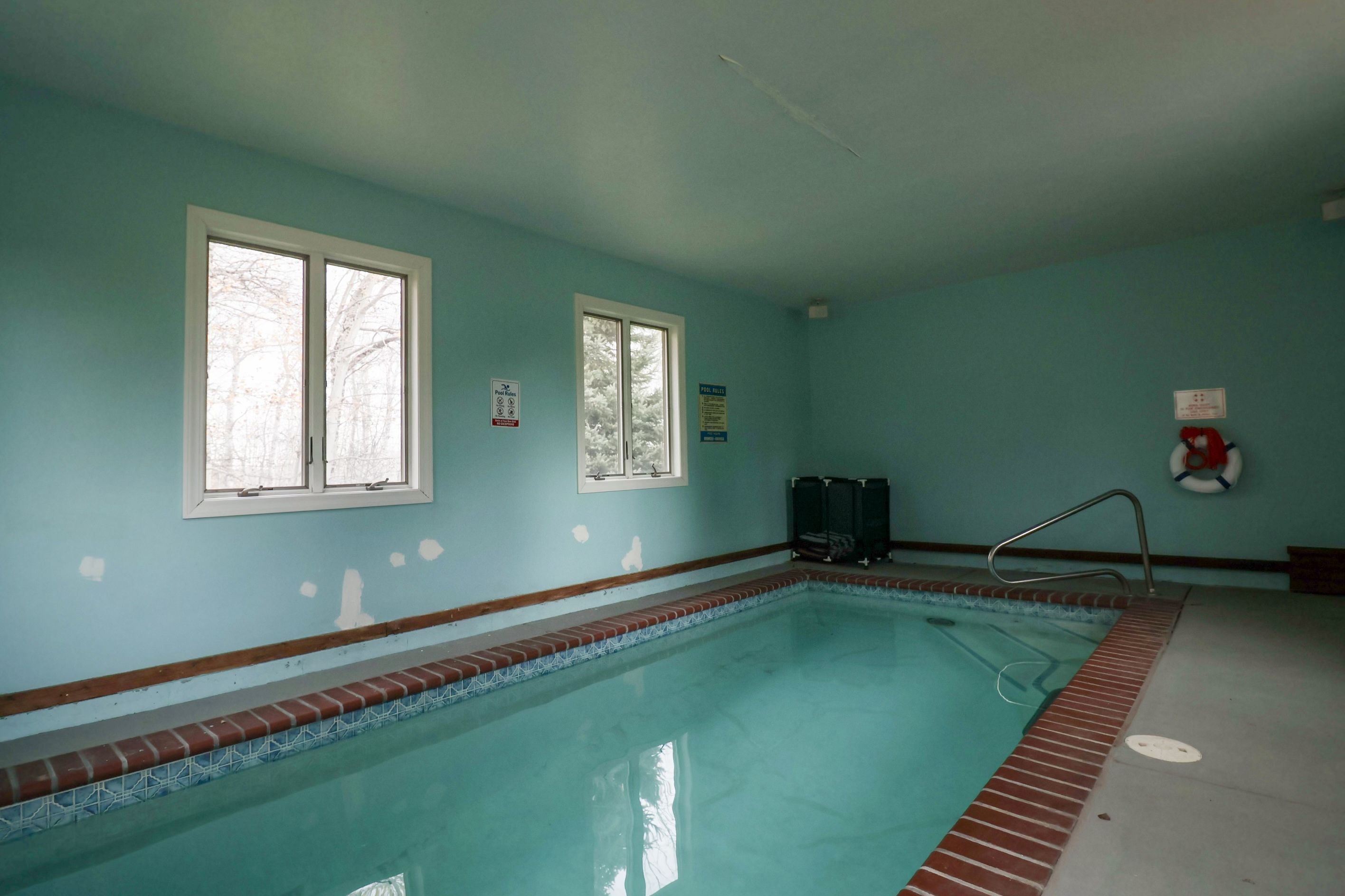 An indoor swimming pool with clear pool rules posted on the walls, a lifebuoy, and a window with a view of trees.