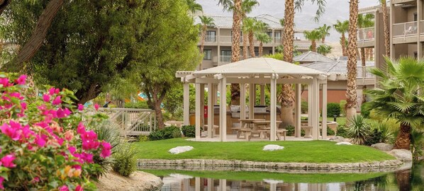 Ein Gazebo mit Picknicktisch in einem angelegten Garten mit Teich und Palmen.