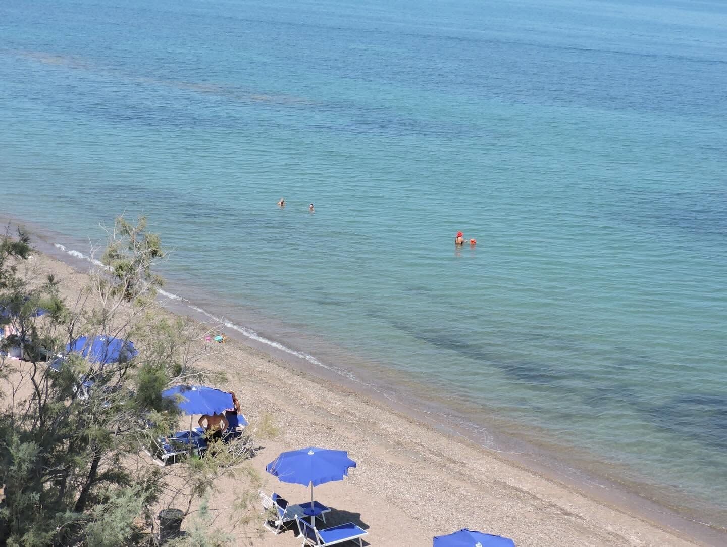 Uma praia com águas cristalinas, algumas pessoas nadando e vários guarda-sóis azuis oferecendo sombra.