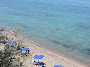 A beach with clear water, a few people swimming, and several blue umbrellas providing shade.