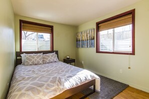 Ein Schlafzimmer mit einem Bett, zwei Fenstern mit Jalousien, einem Nachttisch und einem Bild an der Wand.