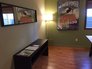 A room with a wooden floor, a bench, a table, a wall-mounted mirror, and two posters advertising ski fresh powder and Aspen Colorado. - Contemporary and Blinged-Out Private Guest Suite (Greeley)