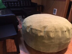 Ein runder, beiger Hocker in einem Raum mit Holzboden, einer schwarzen Ledersofa und einem kleinen Tisch mit einer Fernbedienung.