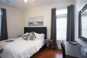 Een hotelkamer met een bed, nachtkastje en een bureau met stoel. Aan de muur hangt een schilderij en er is een raam met gordijnen.