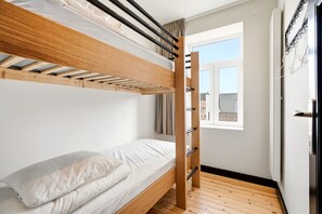 Uma cama de solteiro com estrutura de madeira e roupa de cama branca, uma janela com vista para edifícios e uma porta com puxador de metal.