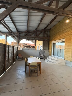 A covered outdoor area with a wooden table and chairs, a hanging chair, and a view of buildings in the background. - House 'Maison 10 personnes grande terrasse grands groupes' (Parcé)