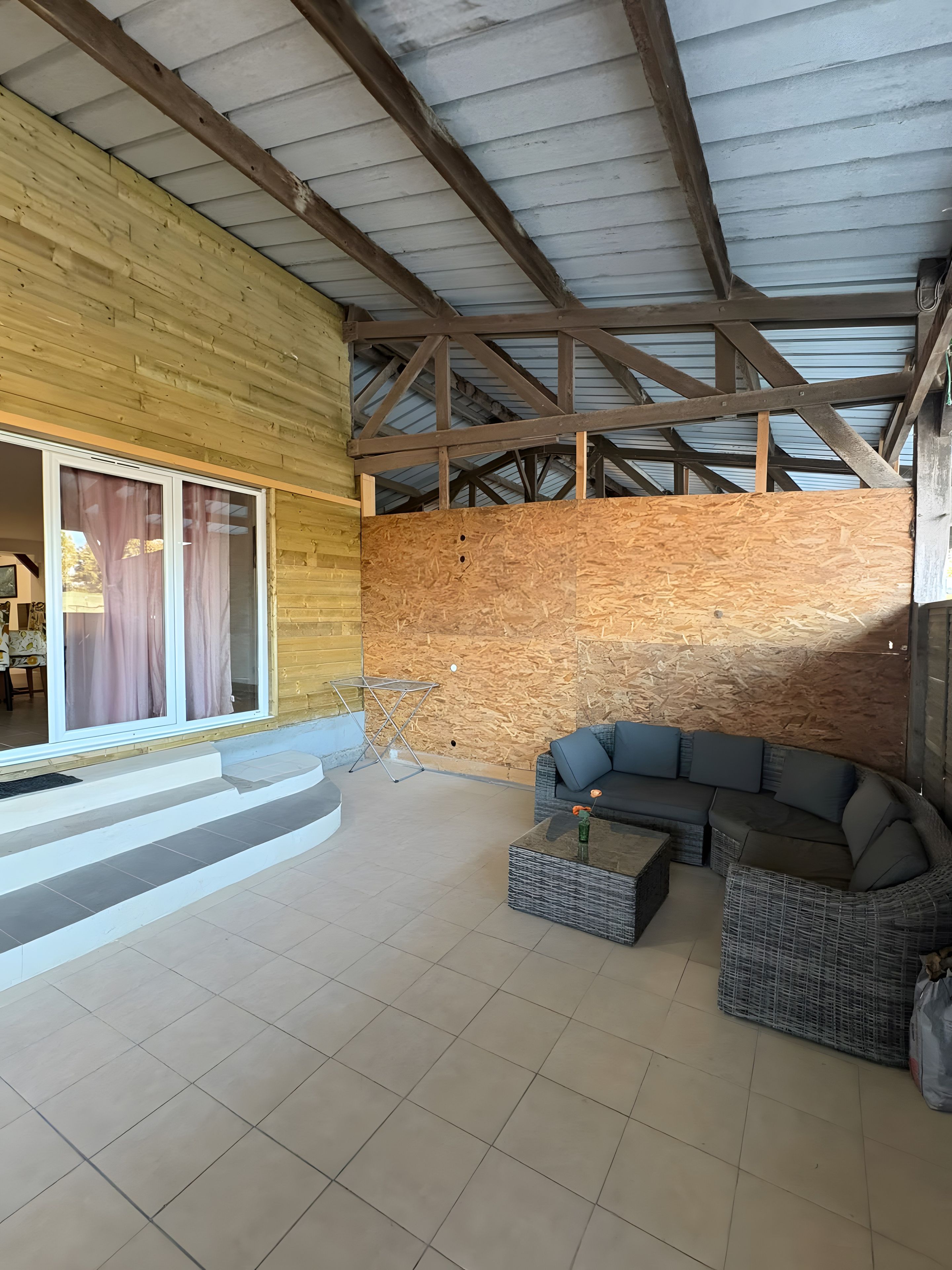 A covered outdoor area with a wooden wall, a sofa, and a glass table.