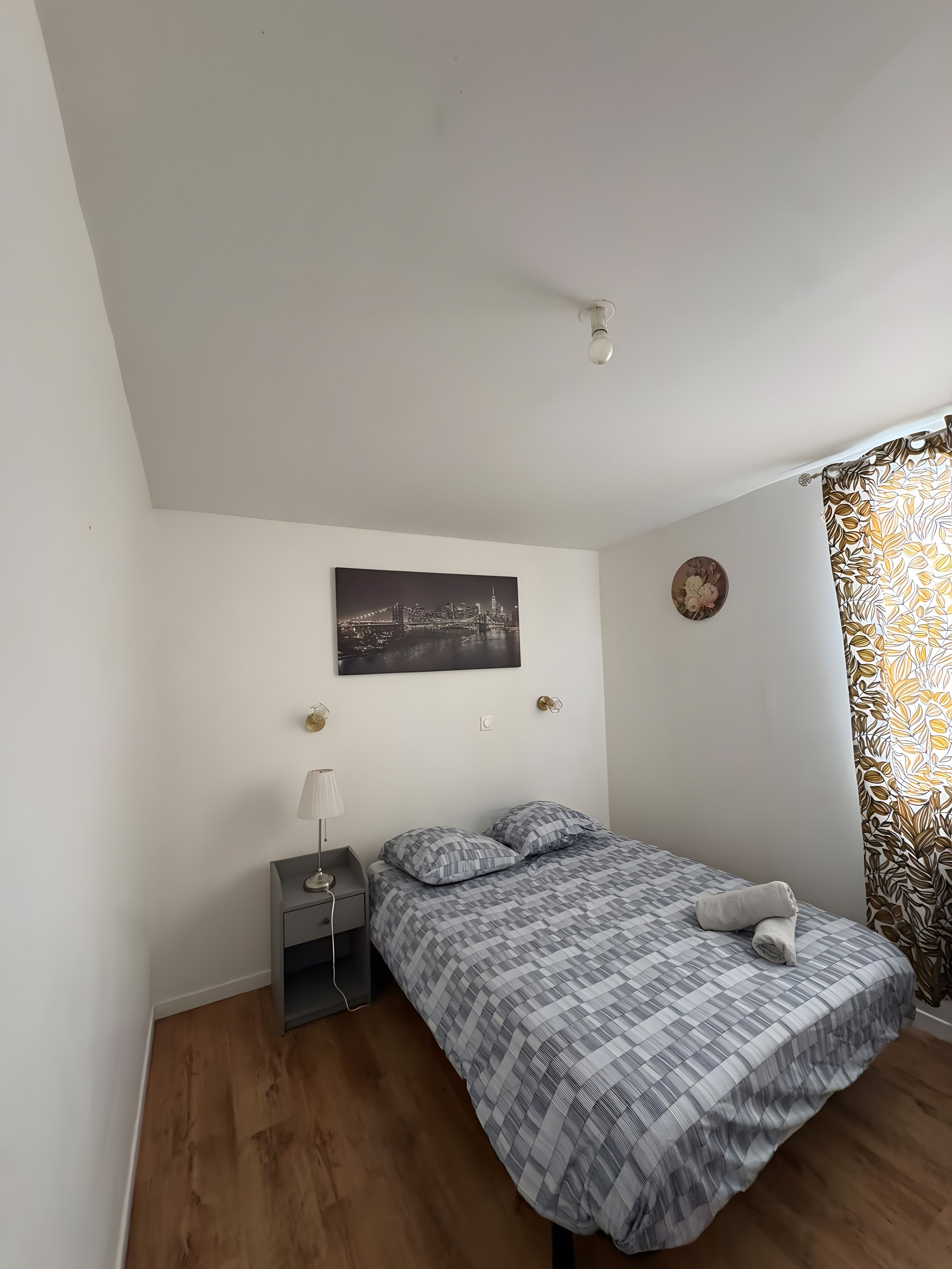 Une chambre à coucher comprenant un lit, une table de chevet, une lampe, un tableau représentant un paysage urbain, une horloge murale et un rideau à motifs.