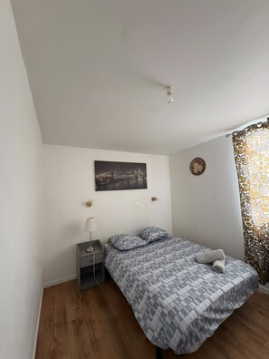 Ein Schlafzimmer mit einem Bett, einem Nachttisch, einer Lampe, einem Stadtbild als Gemälde, einer Wanduhr und einem gemusterten Vorhang.