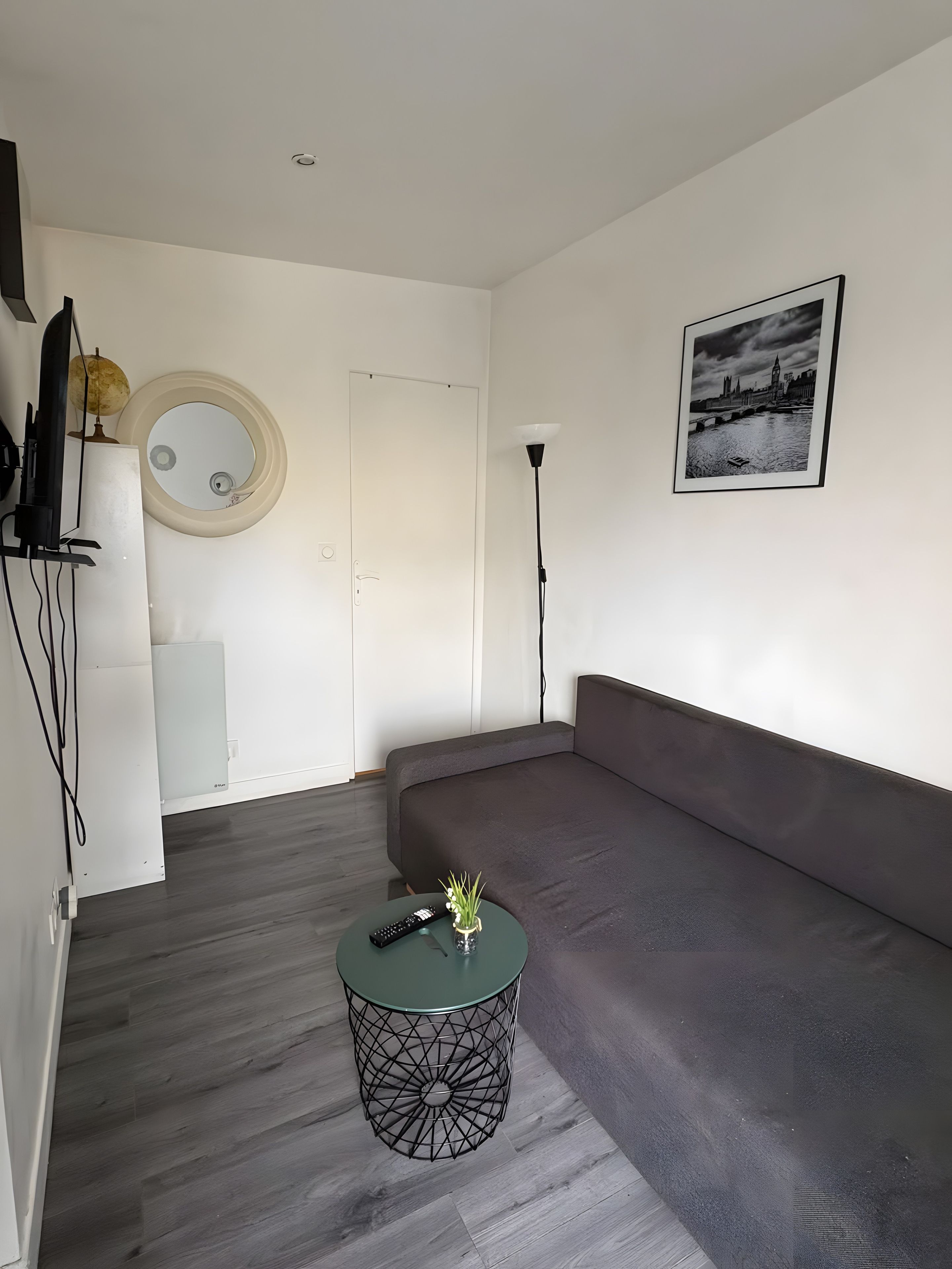 Een moderne woonkamer met een grijze bank, een ronde spiegel, een lijst met een afbeelding en een klein rond tafeltje met een afstandsbediening.