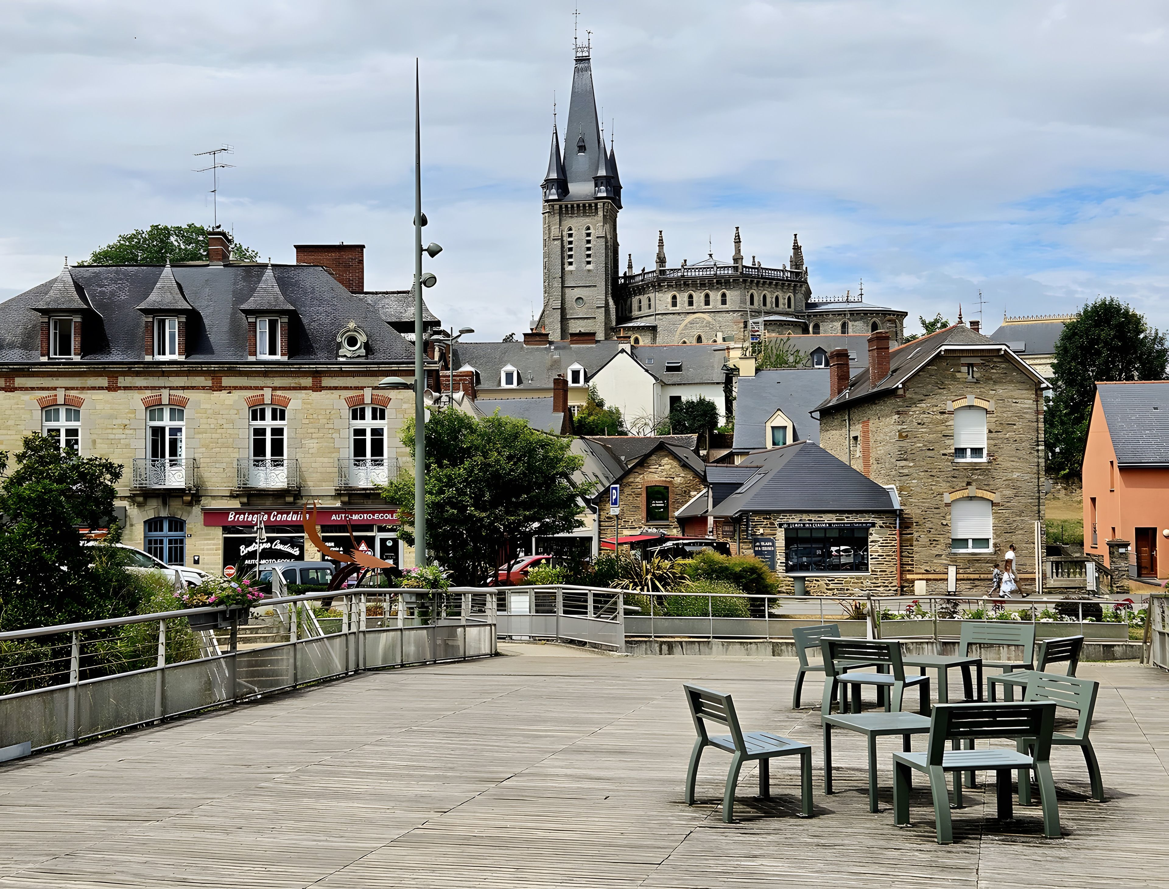 Une place de ville avec des bancs et des tables, des bâtiments historiques et un clocher d’église imposant.