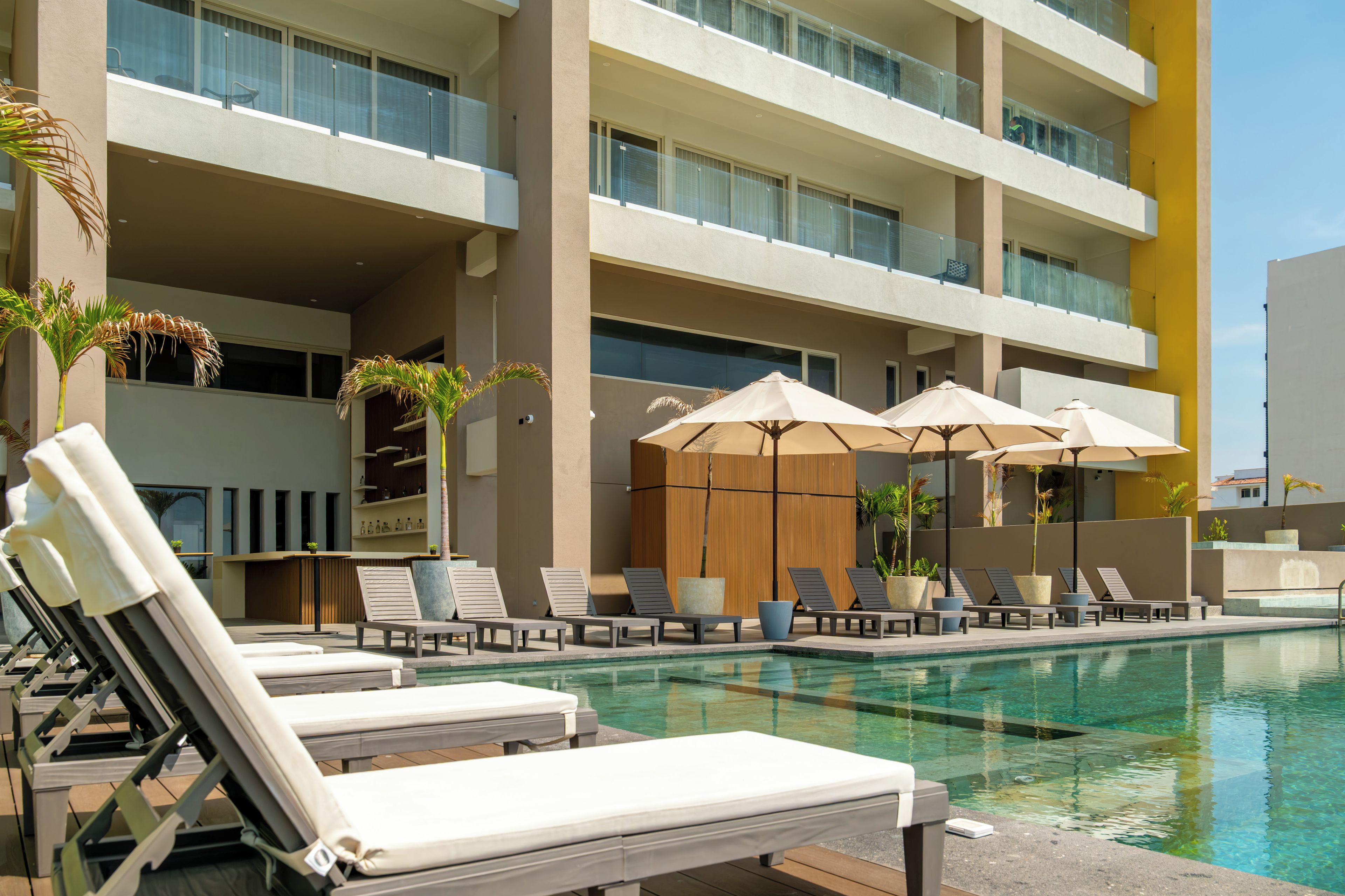 A poolside area with lounge chairs and umbrellas, a wooden fence, and a multi-story building with balconies.