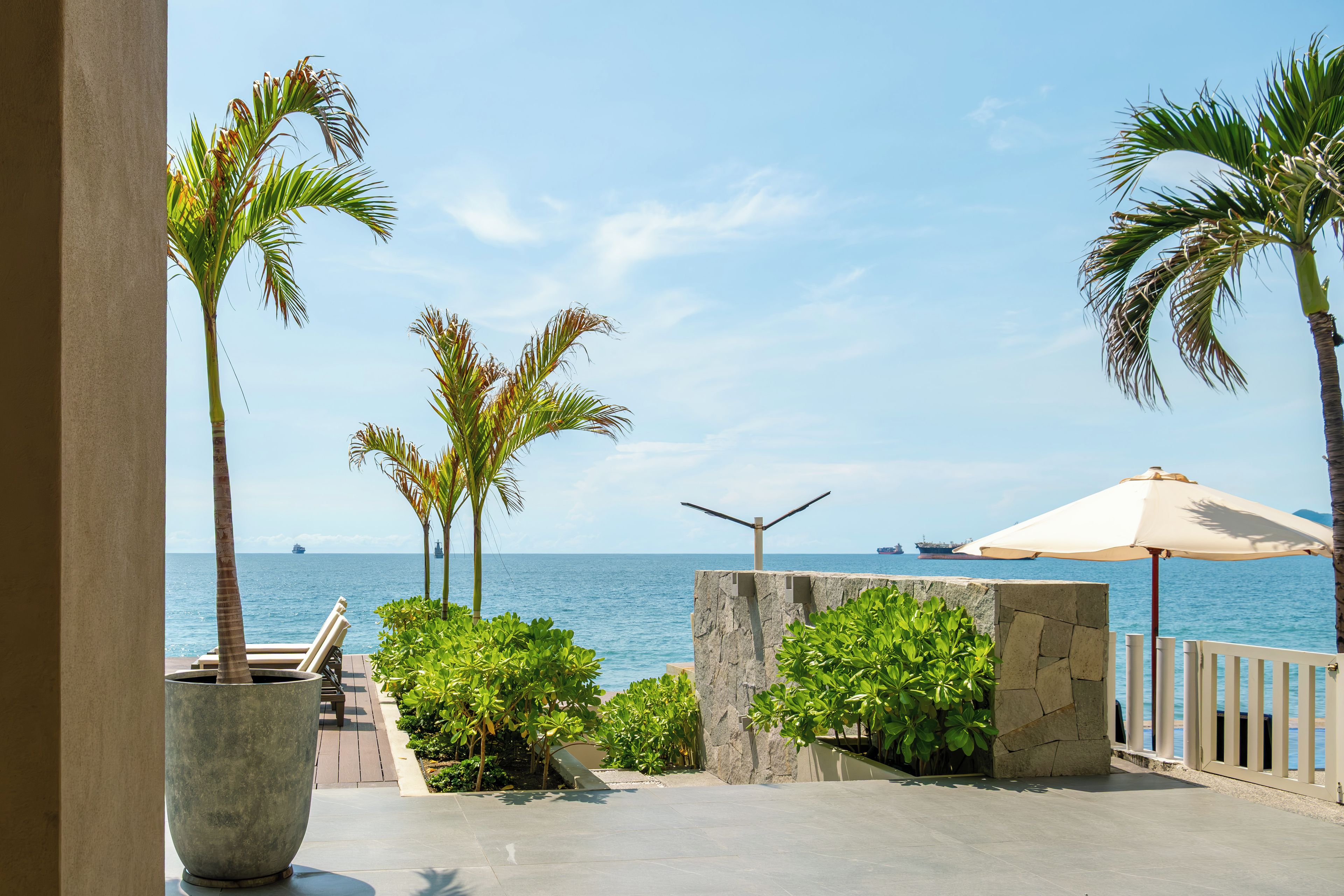 A seaside patio with palm trees, a stone wall, and a view of the ocean.