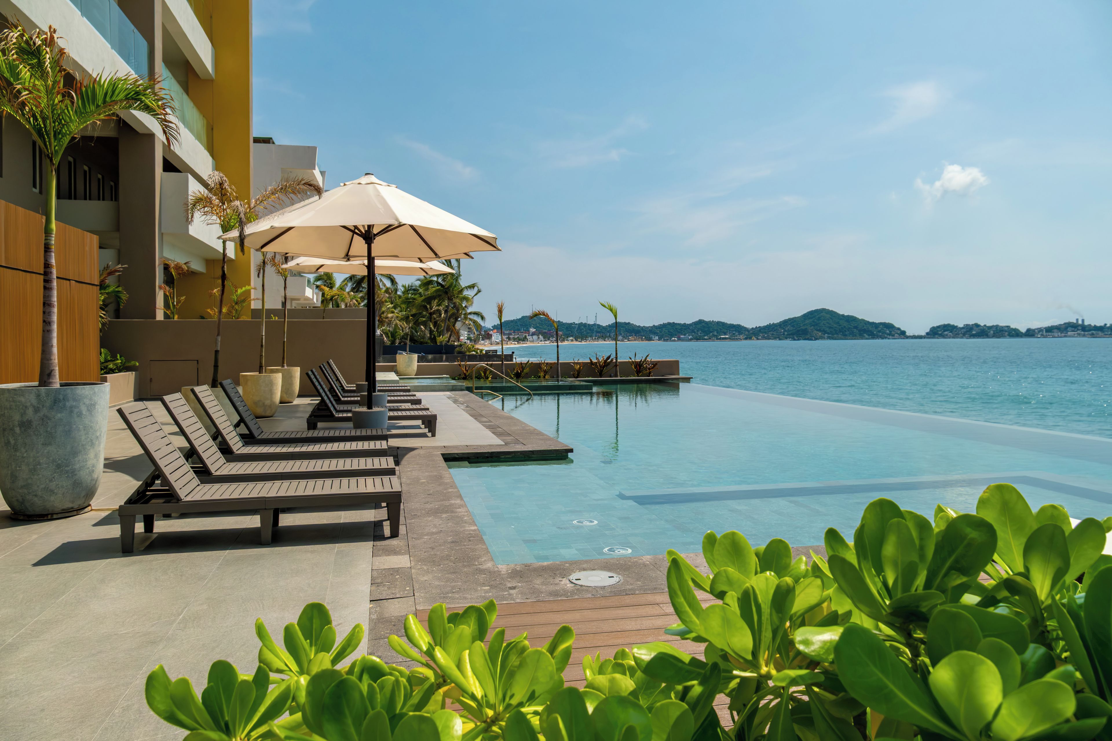 A pool area with lounge chairs and umbrellas, a clear view of the sea, and a distant mountain range.