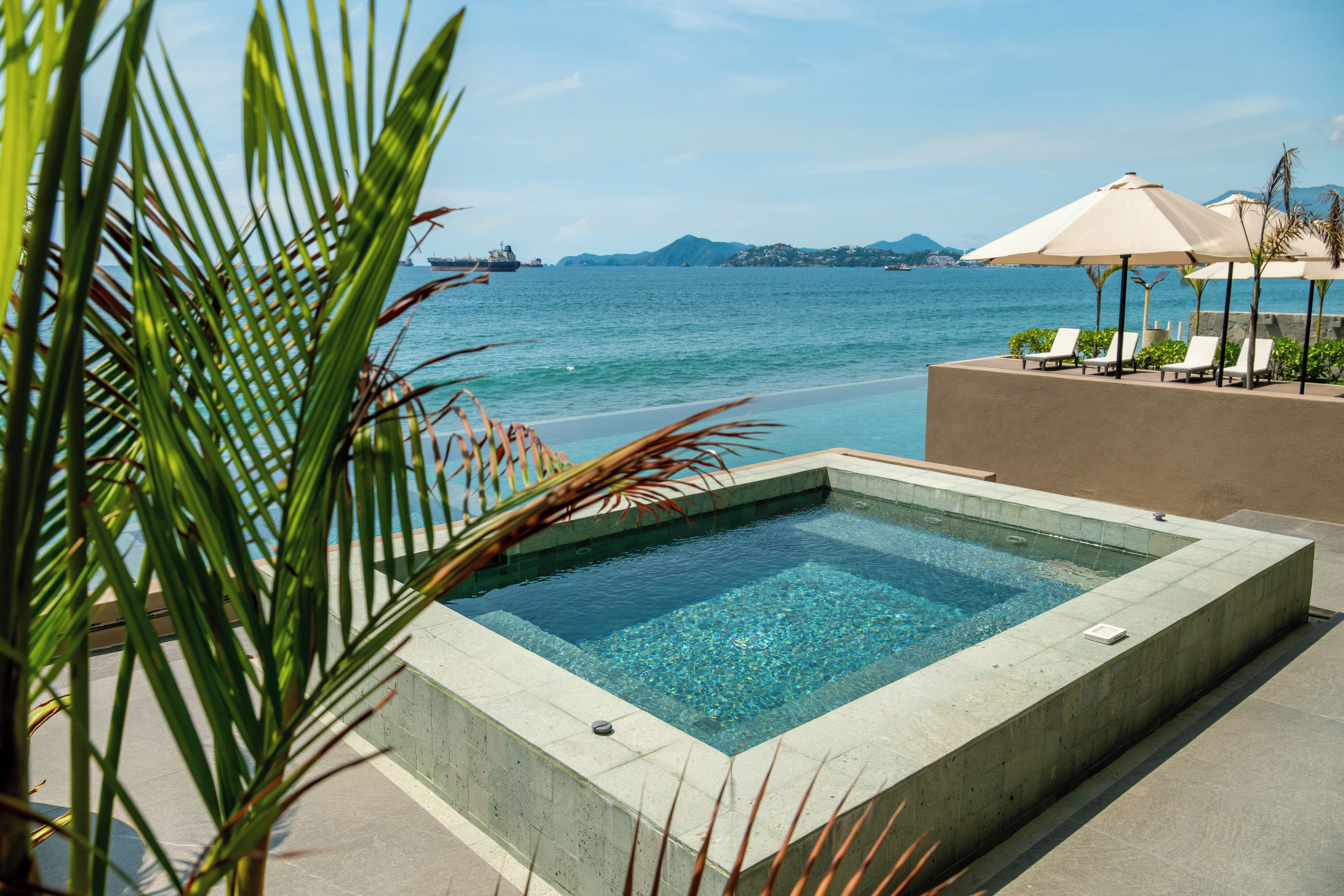 A rectangular infinity pool with clear blue water, surrounded by a concrete deck, with a view of the sea and distant mountains.