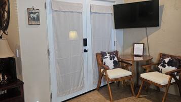 A room with a closed door, a mounted television, two wooden chairs with cushions, and a small table.
