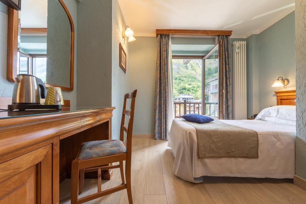 A hotel room with a bed, a desk with a chair, a window with curtains, and a view of greenery outside.