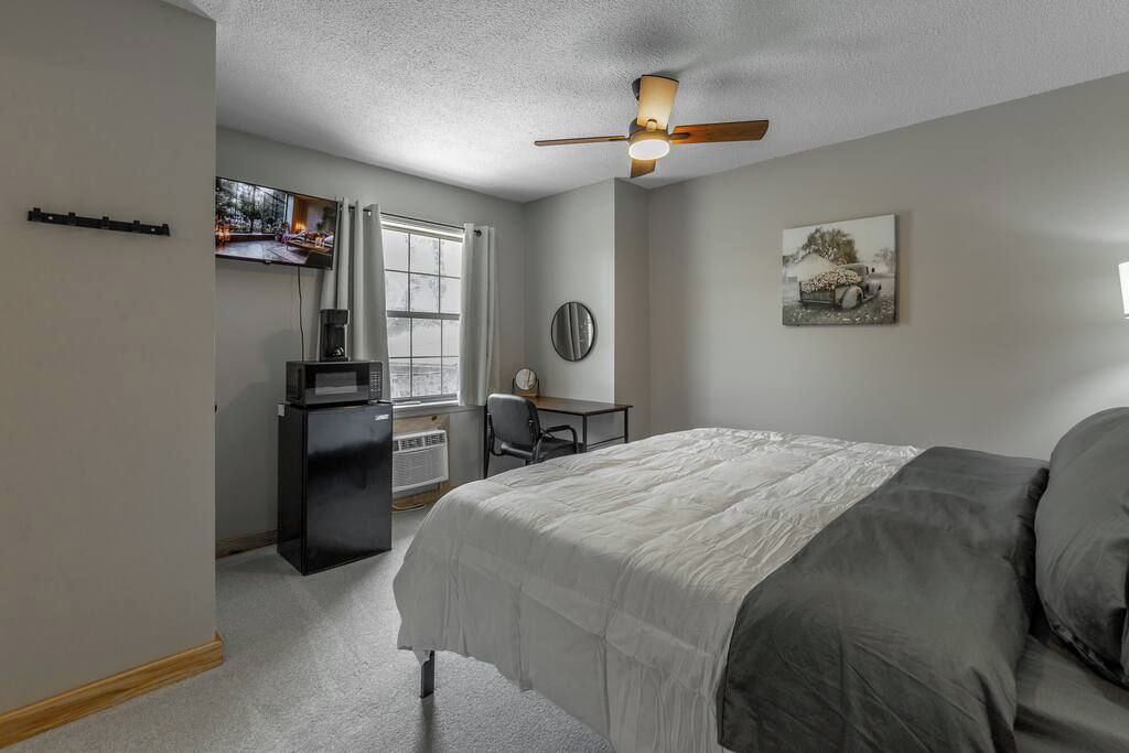 Un dormitorio con cama, escritorio, silla, ventilador de techo y ventana.