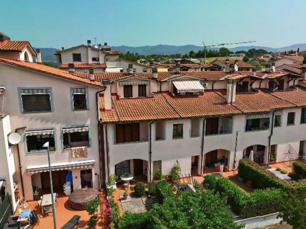 Zona residencial com telhados de telha de barro, um pátio com vegetação e montanhas ao fundo.