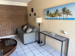 Ein Wohnzimmer mit Backsteinwand, einem grauen Sofa, einer Stehlampe, einem kleinen Tisch und einem Palmenbild.