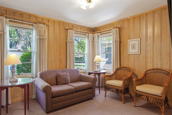 A room with a brown sofa, two wicker chairs, a small table with a lamp, and a framed picture on the wall.