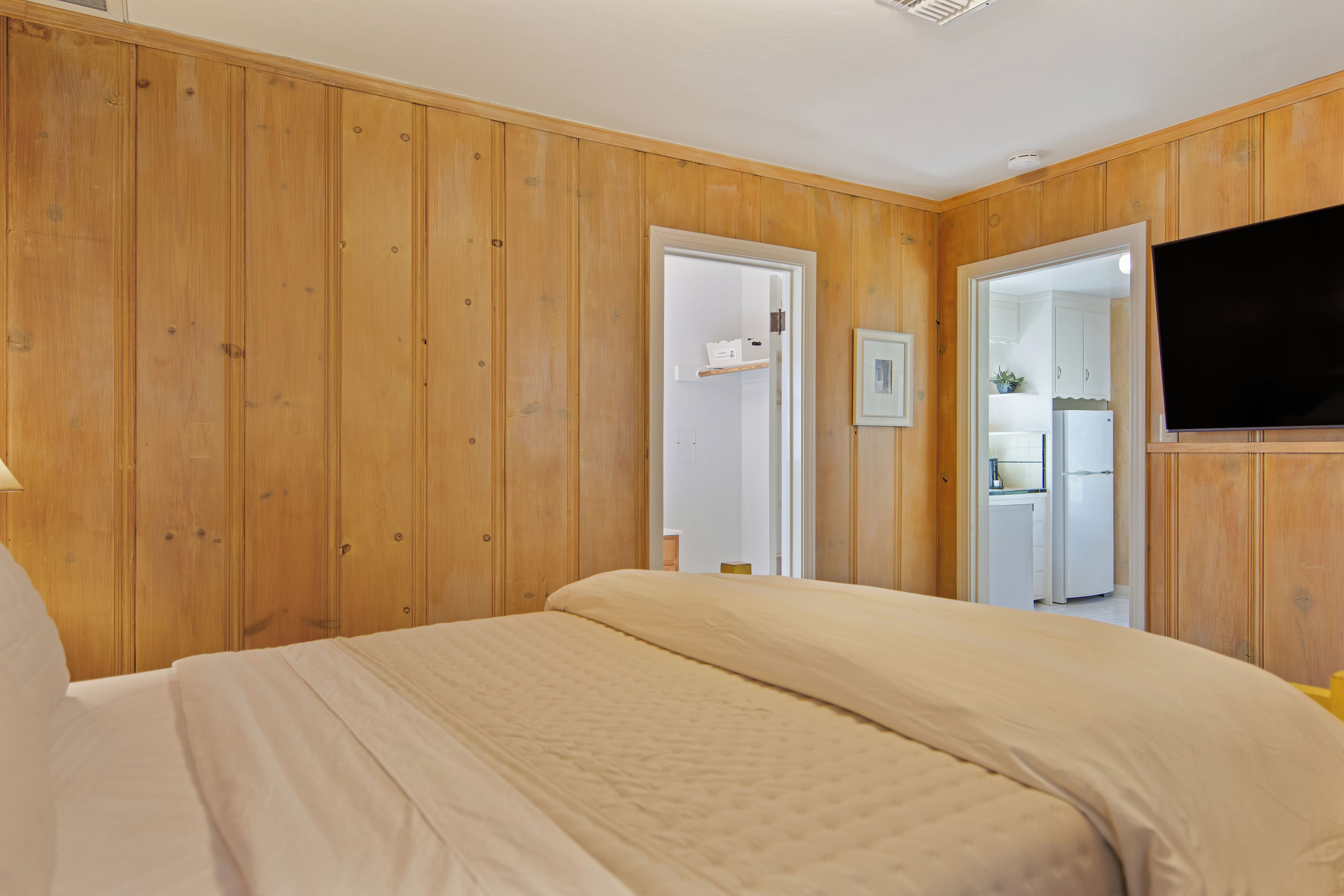 A bedroom with wooden paneling, a bed with a beige duvet, a television on the wall, and a door leading to a bathroom.