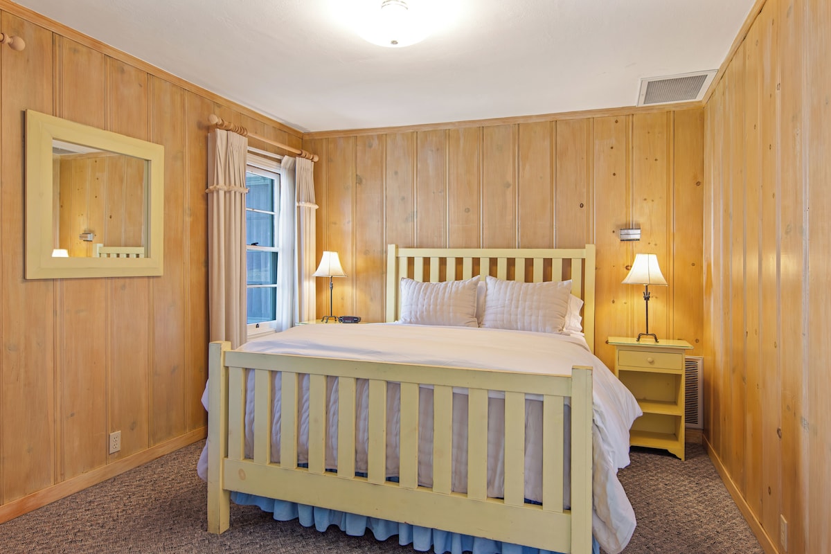 Un dormitorio con una cama de madera, dos mesitas de noche con lámparas, una ventana con cortinas y un espejo colgado en la pared.