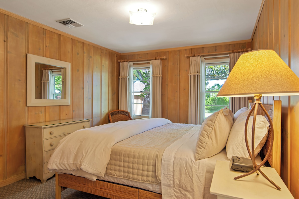 Un dormitorio con cama, mesita de noche, lámpara y ventana con cortinas.