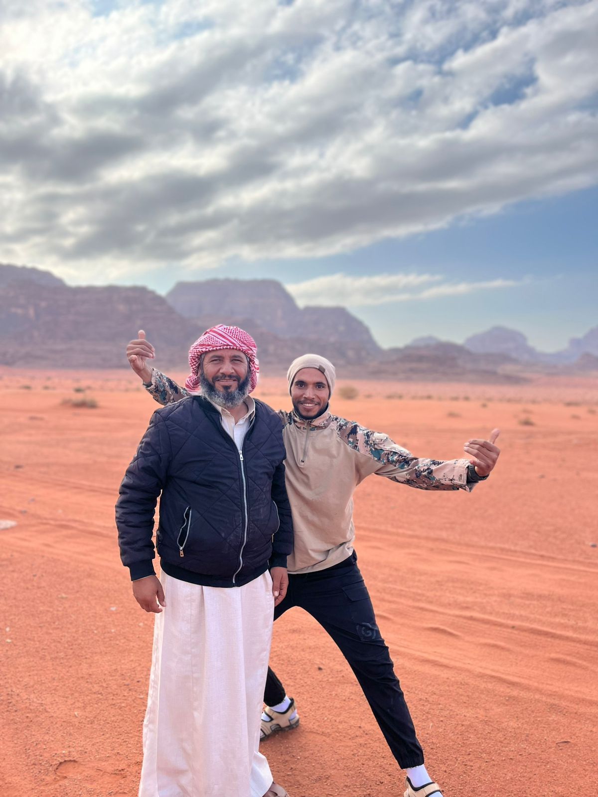 Two individuals in a desert landscape with mountains in the background.