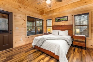 A wooden cabin bedroom with a bed, nightstands, and large windows. - Bear Mountain Hideaway~Theater Room~Game Room~Hot-Tub (Sevierville)