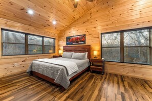 A wooden cabin bedroom with a bed, two bedside tables, a nightstand, and a painting on the wall. - Bear Mountain Hideaway~Theater Room~Game Room~Hot-Tub (Sevierville)