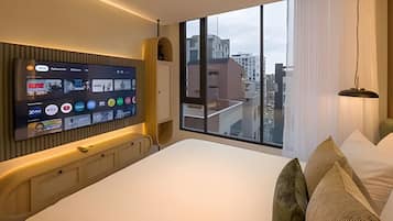 Habitación de hotel moderna con una cama grande, un televisor montado en la pared y vistas de la ciudad a través de un ventanal.