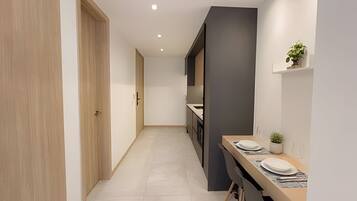 A narrow hallway with a wooden door on the left, a kitchen area with a countertop and cabinets on the right, and a small shelf with a plant.