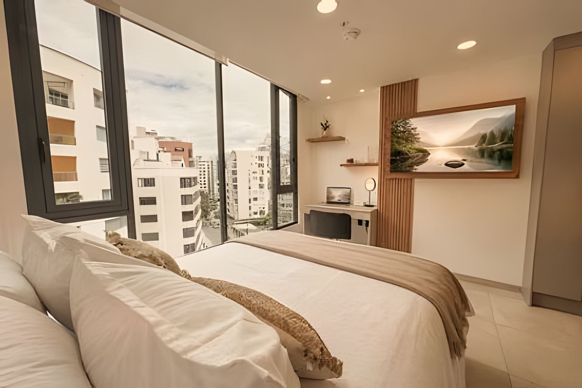 A modern bedroom with a large bed, a desk, and a view of the city through large windows.
