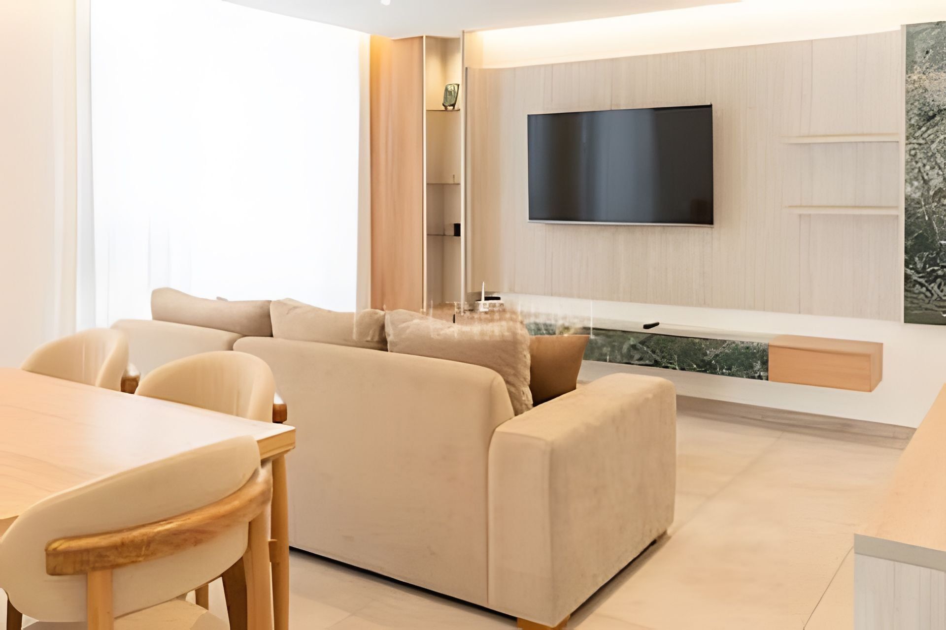 A modern living room with a sofa, a dining table, and chairs. A flat-screen TV is mounted on the wall.