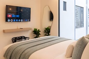 Una habitació d'hotel moderna amb un llit, una televisió penjada a la pared, una taula de nit i una finestra amb vista a edificis.