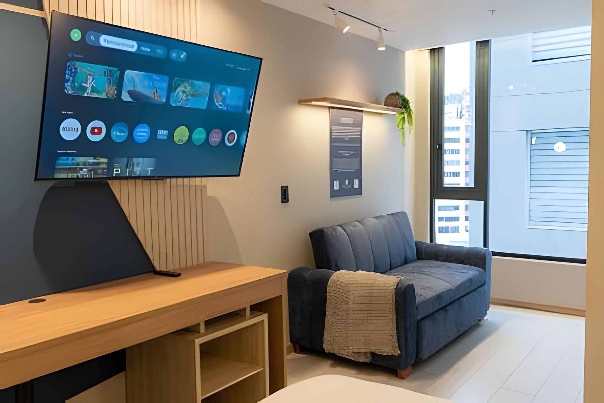 A modern living room with a flat-screen TV mounted on the wall, a wooden entertainment unit, a blue sofa, and a large window providing a view of the city.