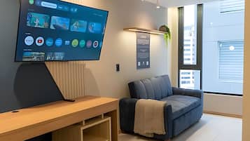 A modern living room with a flat-screen TV mounted on the wall, a wooden entertainment unit, a blue sofa, and a large window providing a view of the city.