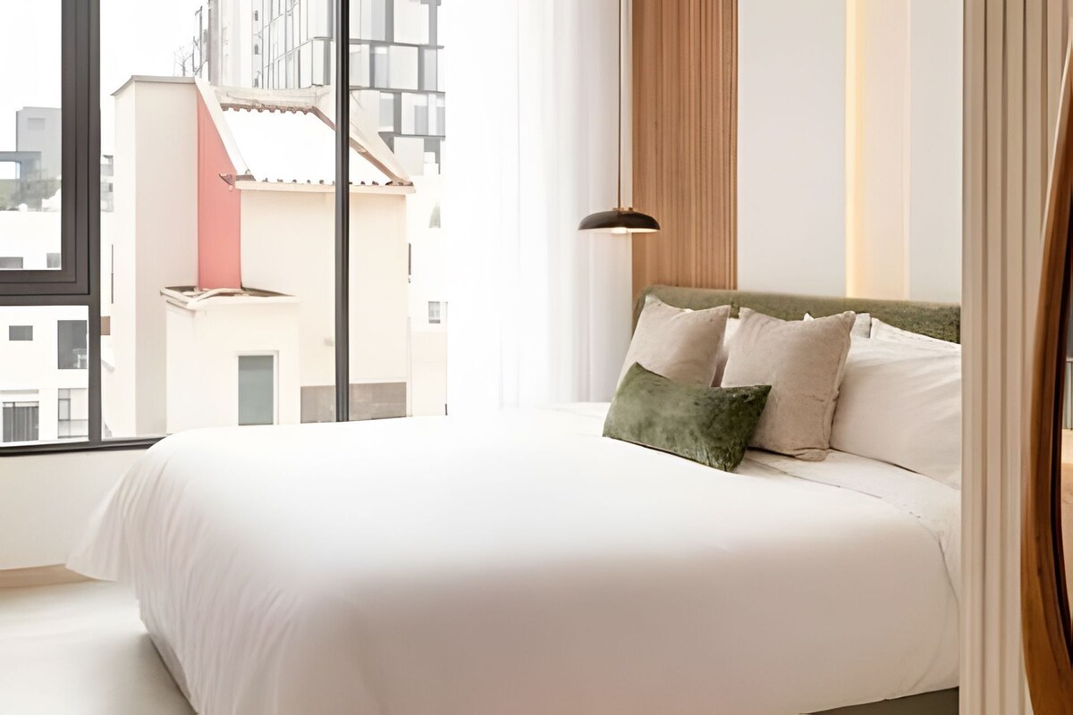 Habitación de hotel con una cama grande, vistas de edificios a través de la ventana y una lámpara colgante moderna.