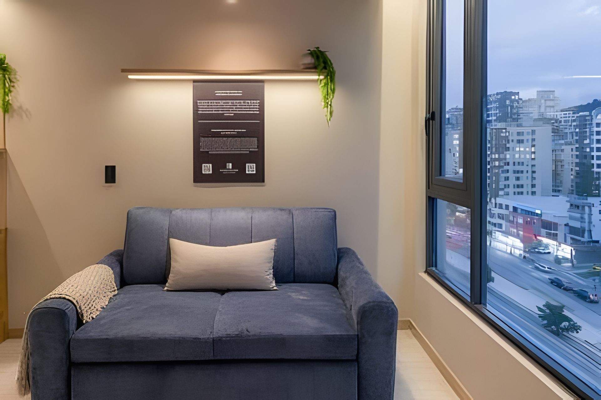 Een moderne woonkamer met een grijze bank, een kleine plant en een stadszicht door grote ramen.