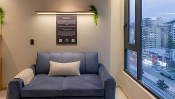 Een moderne woonkamer met een grijze bank, een kleine plant en een stadszicht door grote ramen.