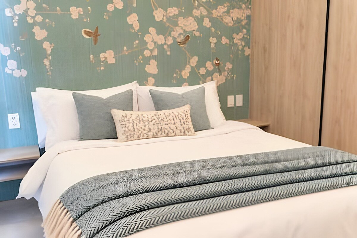 Una cama con una colcha y almohadas estampadas, una mesita de noche de madera y una pared con papel tapiz floral.