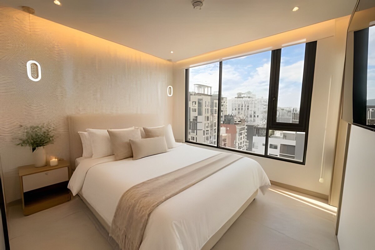 Un dormitorio moderno con una cama grande, una mesita de noche con una planta y vistas a la ciudad a través de amplias ventanas.