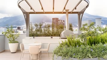 A rooftop terrace with a covered seating area, potted plants, and a view of a cityscape and mountains.